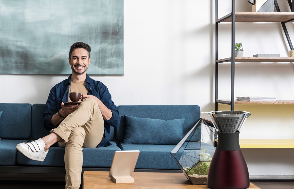 Portrait of happy young man with beard sitting cross-legged on cozy couch and drinking coffee. He is holding cup of coffee in his hands and smiling