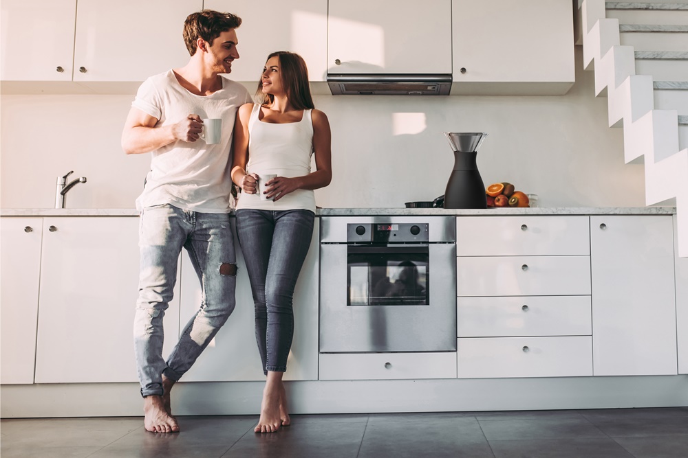 Full-length image of romantic couple at home. Attractive young woman and handsome man are enjoying spending time together while standing on light modern kitchen with cup of coffee in hands.